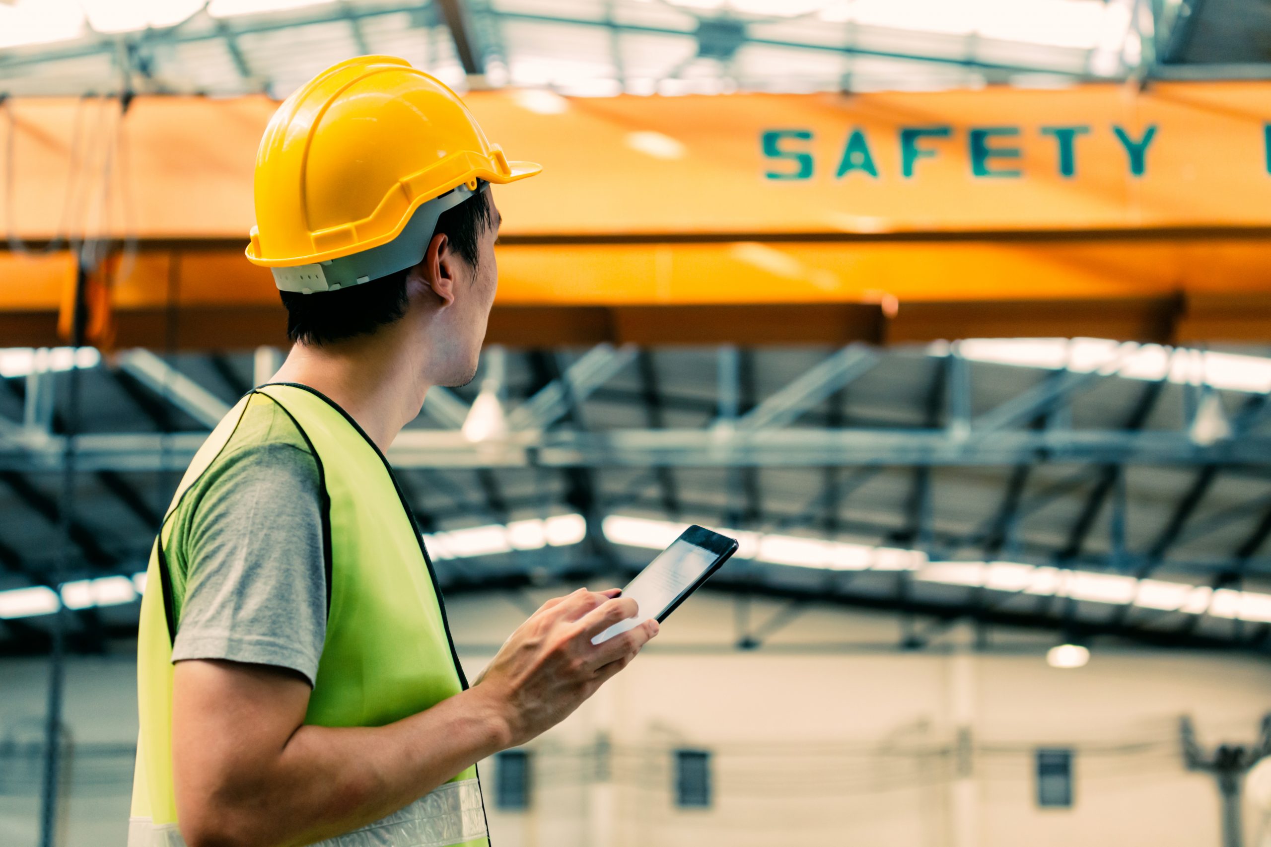 Young Asian male heavy industral worker using a digital tablet inside manufacturing and factory site with crane behind